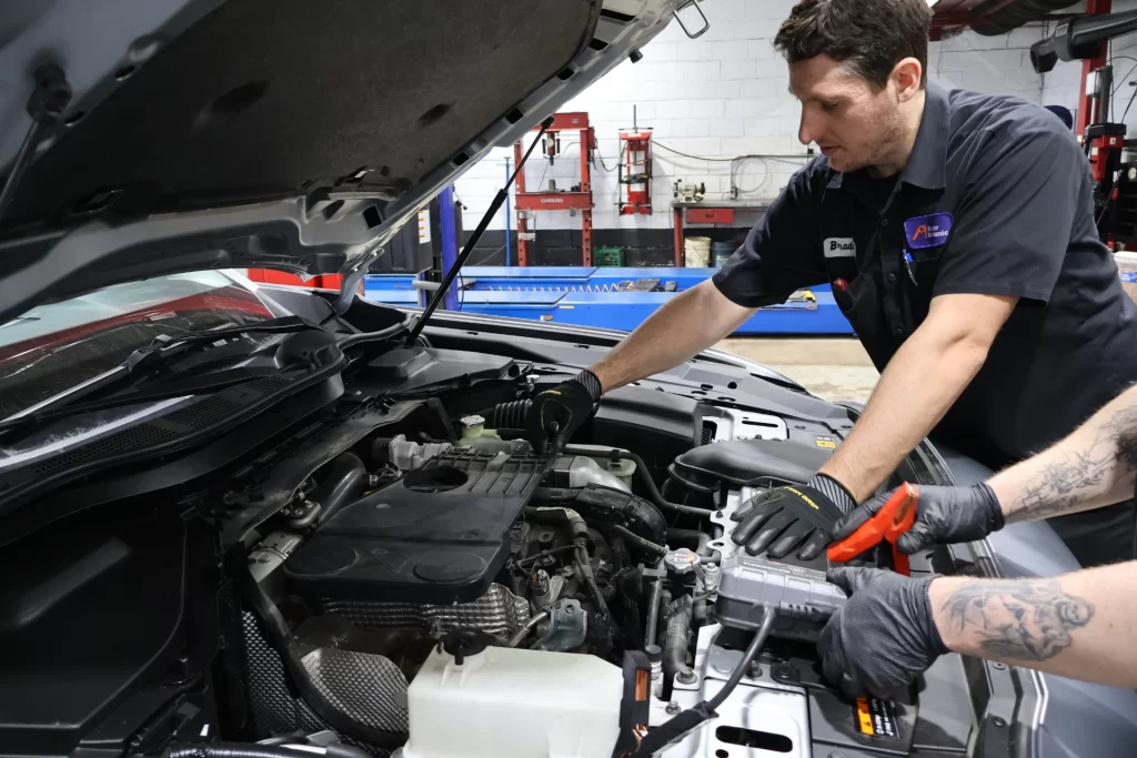Two Master Mechanic technicians work under the hood of a car in a garage, one inspecting the engine while the other uses a handheld diagnostic tool